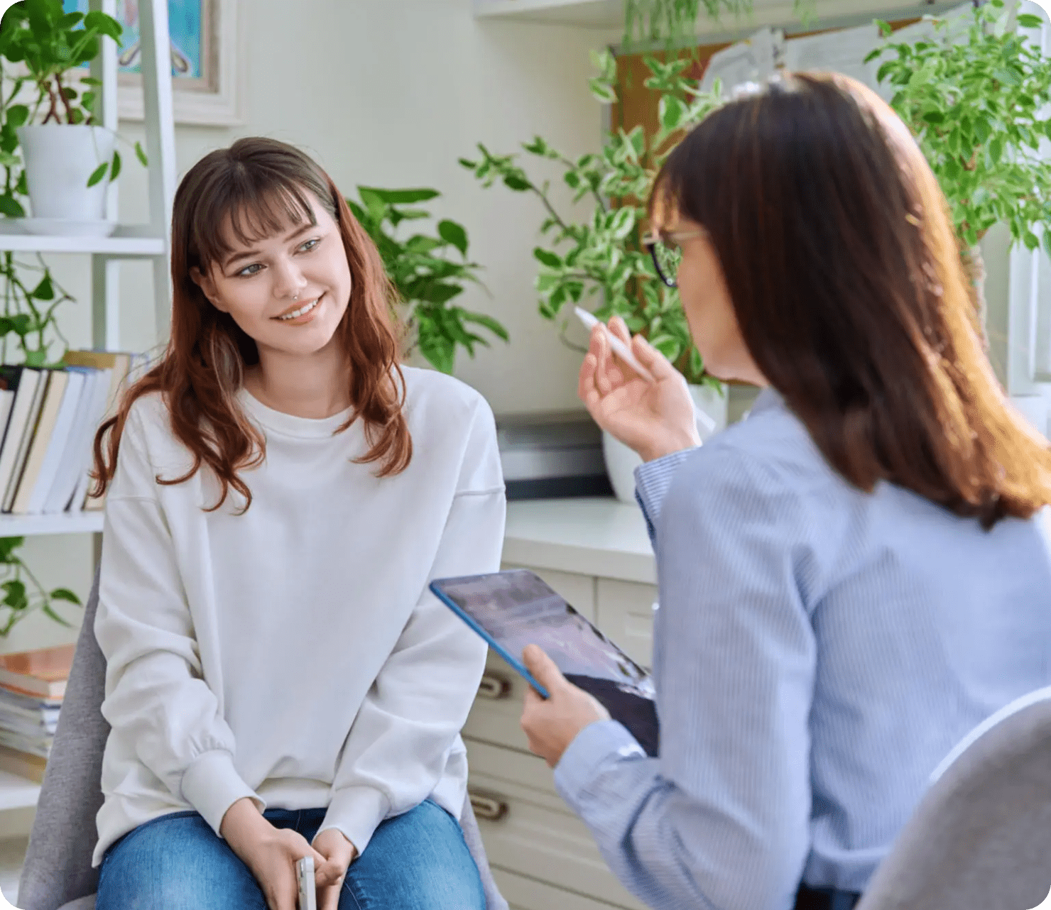 Two women having a discussion in a bright room filled with plants and books, one holding a tablet and a pen.
