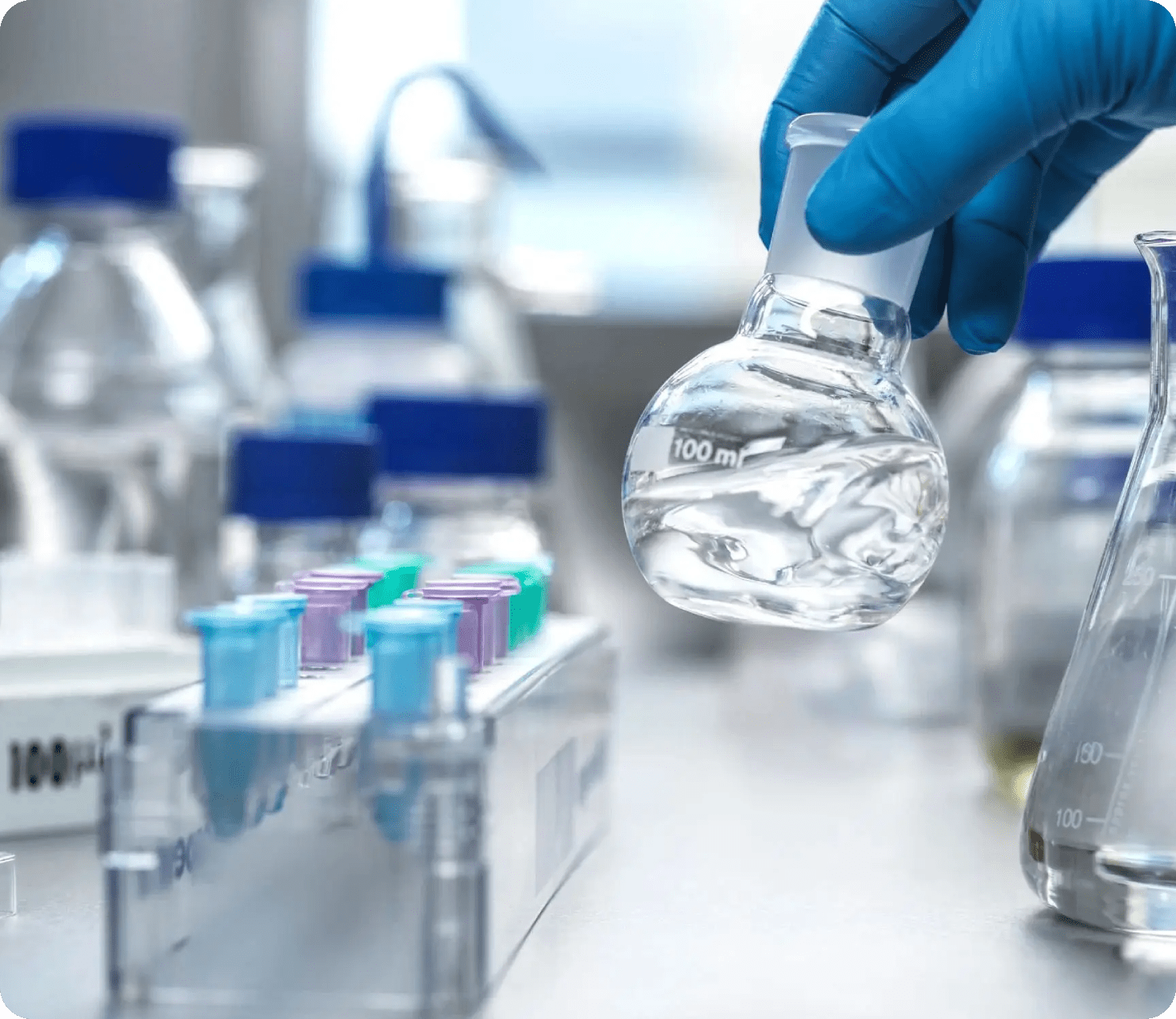 A laboratory scene featuring a hand holding a flask with liquid, surrounded by various test tubes and bottles on a table.