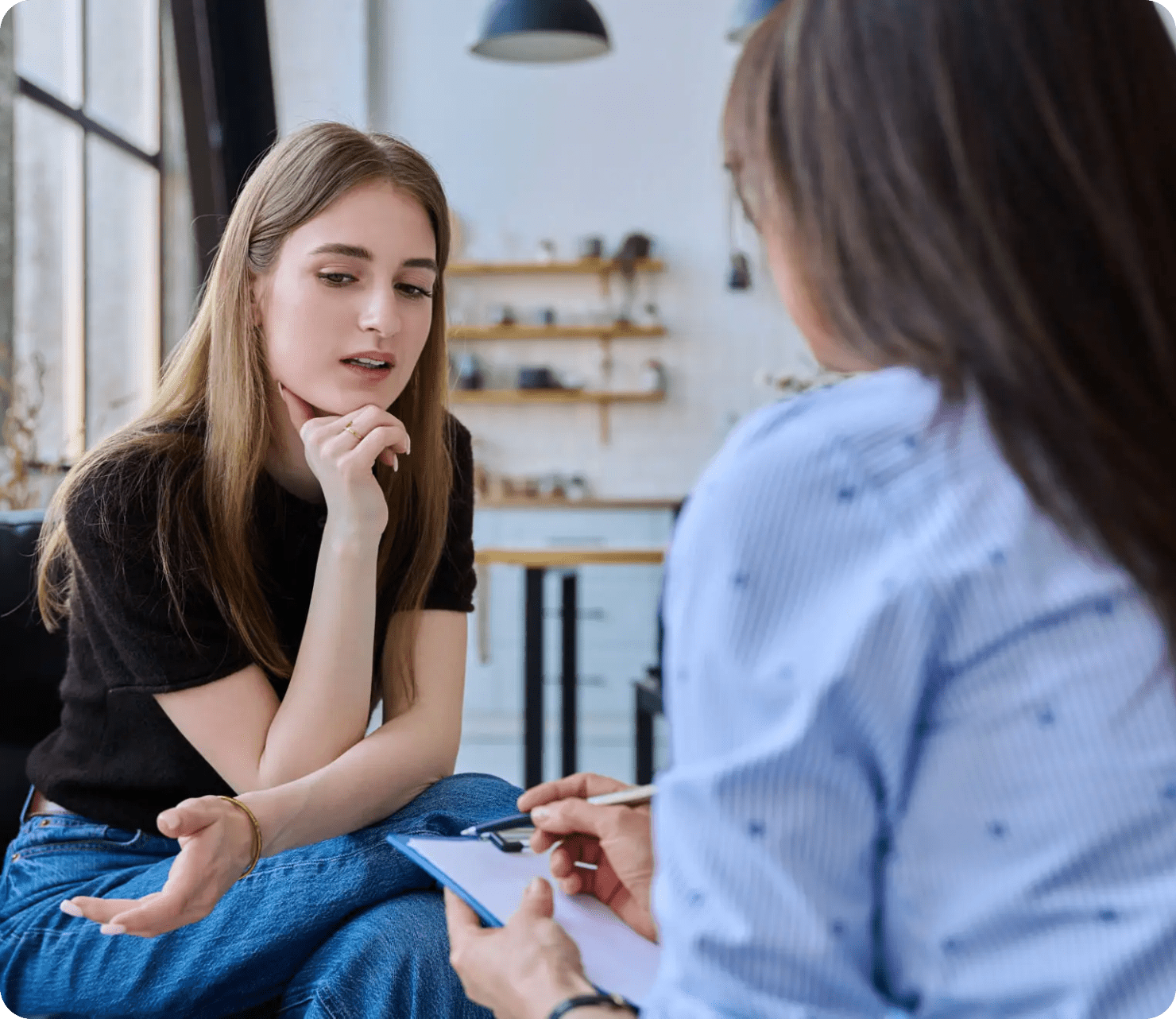 Two women in a counseling session, one talking and the other taking notes on a clipboard in a bright, modern room.